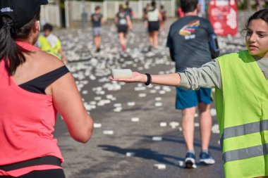 Nigran, Pontevedra, İspanya; 6 Nisan 2025: Yüksek görüşlü yelek giyen genç gönüllüler İspanya 'nın Nigran kentinde düzenlenen Vig Bay Half Marathon sırasında koşuculara suya batırılmış sünger dağıtıyorlar. Sahne, toplum desteği ve ekip çalışmasının bir anını partik olarak yakalıyor.