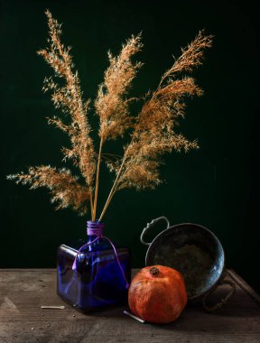 Mavi cam bir vazoda kurutulmuş bir buket bitki, olgun bir nar ve koyu arkaplanda bakır kaplarla dolu bir hayat. Eski.
