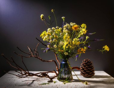 Cam vazodaki masanın üzerinde bir buket çayır çiçeği. Yakınlarda bir dal ve büyük bir çam kozalağı var. Bir ışık demeti çiçekleri aydınlatır..