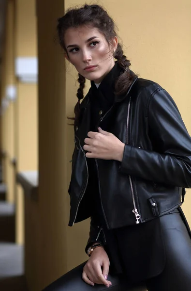 革のジャケットにピグテールとベージュの壁に対する黒のレギンスを持つ美しい女の子の肖像画 — ストック写真