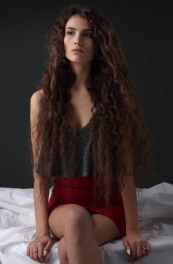 Uzun, kalın saçlı, parlak bluzlu ve koyu arkaplanda kırmızı etekli bir kız.