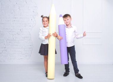 Beyaz arka planda büyük kalemler tutan okul üniforması giymiş bir erkek ve bir kız.