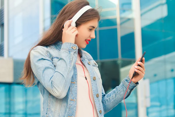 Красивая девушка слушает музыку в наушниках и улыбается глядя на телефон — стоковое фото