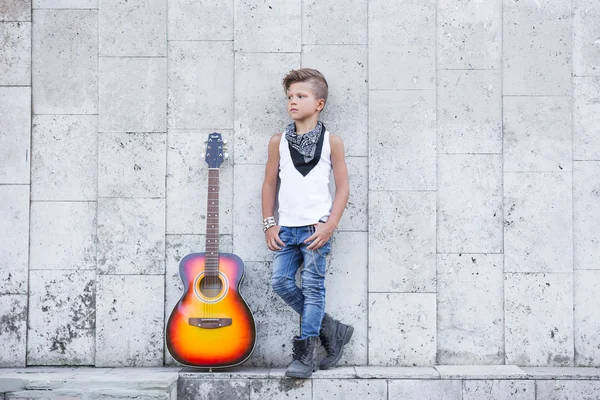 A little Boy stands next to a guitar and put his hands in his pocket