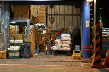 Tayvan - 19 Ekim 2025: Gece açık depo girişi istiflenmiş karton kutular, el arabası beyaz çantalar, bisiklet, mavi sütun, yuvarlanan kepenk ve ışığı yansıtan ıslak kaldırım.