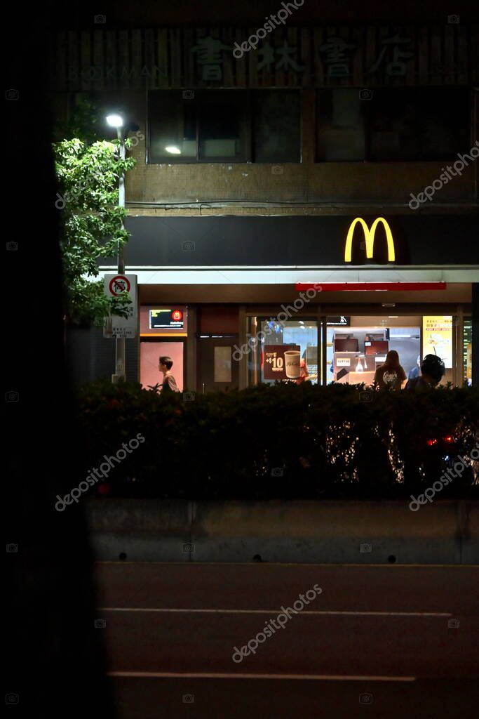 Taipei, Taiwan - Sep.  29, 2025: McDonald's restaurant with yellow M logo glowing at night, customers dining inside by windows, green landscaping and street lamp lighting tree in foreground outdoors.