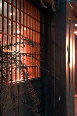 Ön planda kurumuş bitki dallarının arkasında parlak turuncu-kırmızı ışıkla parlayan ahşap kafes penceresi, açık havada sıcak atmosferik aydınlatma yaratmanın yanında koyu renk ahşap duvar..