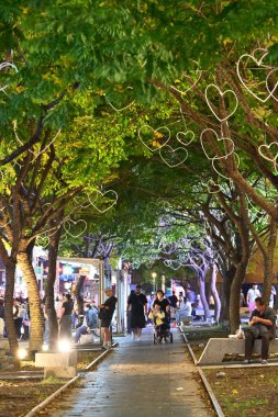 TWN - 26 Ekim 2025: Ağaçlarla süslenmiş tuğla patika, kalp şeklinde ışıklar, yeşil ve sarı yapraklar, gövdelerde aydınlatma, banklarda oturan yayalar ve yürüme mesafeleri, parlak tezgahlar.