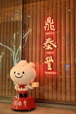 Tayvan - 26 Ekim 2025: Kırmızı üniformalı beyaz çizgi film xiaolongbao robotu siyah pantolon, kırmızı papyon, tepsi, Din Tai Fung yazılı kırmızı tabaka, arkasında ışıklı işaret olan ahşap kapı.