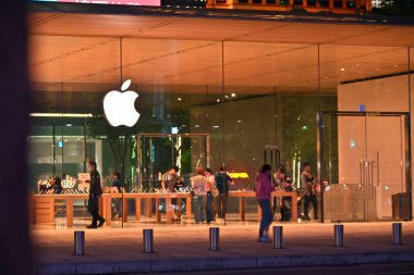 Tayvan - 26 Ekim 2025: Cam cephede parlayan beyaz Apple logosu, içi ahşap masalarla dolu parlak sıcak bir iç mekan, içeride müşteriler ve personel, geceleri dışarıda metal yayan ve yayalar.