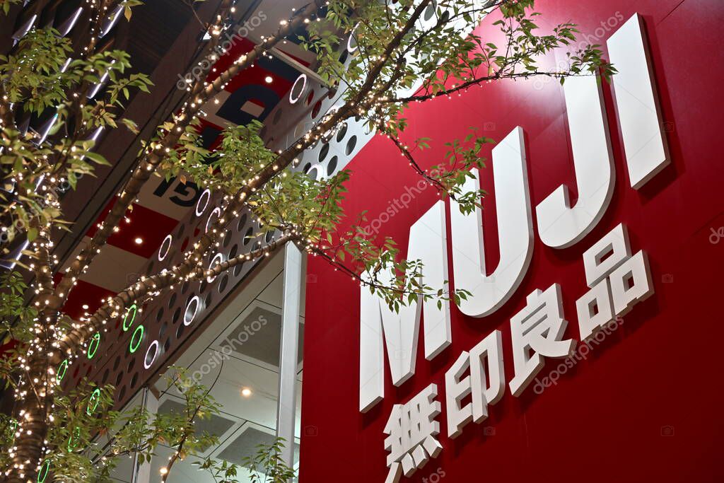 Taiwan - Oct. 26, 2025: Large red sign with white MUJI on building, partially blocked by tree branches and green leaves, illuminated string lights on side, night setting with dark ambient.