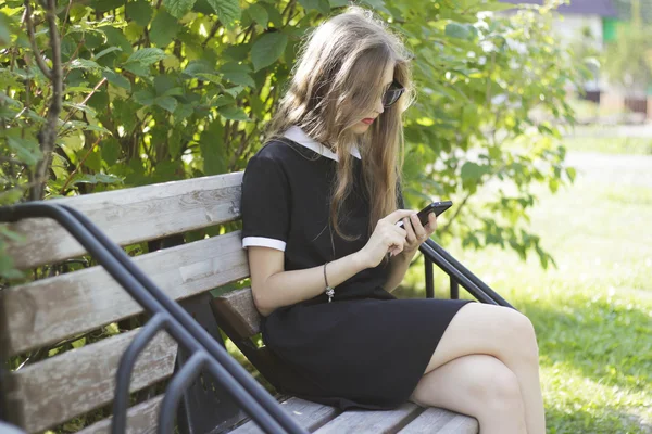 Menina bonita em óculos de sol pretos usar um telefone em um banco de parque — Fotografia de Stock