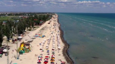 Yazın sahilde güzel bir uçuş. İnsanlar deniz kenarında dinleniyor. Turistler için evler. Plaj şemsiyeleri, gezintiler, denizde yüzen insanlar..