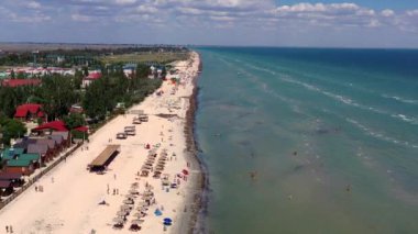 Yazın sahilde güzel bir uçuş. İnsanlar deniz kenarında dinleniyor. Turistler için evler. Plaj şemsiyeleri, gezintiler, denizde yüzen insanlar..