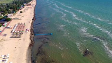 Yazın sahilde güzel bir uçuş. İnsanlar deniz kenarında dinleniyor. Turistler için evler. Plaj şemsiyeleri, gezintiler, denizde yüzen insanlar..