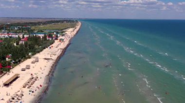 Yazın sahilde güzel bir uçuş. İnsanlar deniz kenarında dinleniyor. Turistler için evler. Plaj şemsiyeleri, gezintiler, denizde yüzen insanlar..