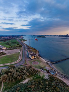 Gün batımında kıyı şeridindeki hava manzarası, helikopter pisti yarımadası, altın sudaki gemi, ufuktaki rüzgar türbinleri ve konut şebekesi deniz aktivitesini yenilenebilir enerji ile harmanlıyor..