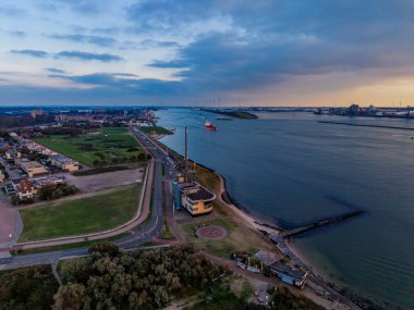 Gün batımında kıyı şeridindeki hava manzarası, helikopter pisti yarımadası, altın sudaki gemi, ufuktaki rüzgar türbinleri ve konut şebekesi deniz aktivitesini yenilenebilir enerji ile harmanlıyor..