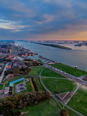 Günbatımı kargo gemileri, vinçler ve endüstriyel rıhtımlardaki nehir limanının havadan görünüşü, yoğun şehir şebekesi ve yeşil alanlar dinamik ticari bir manzarada su yolunu çerçeveliyor..