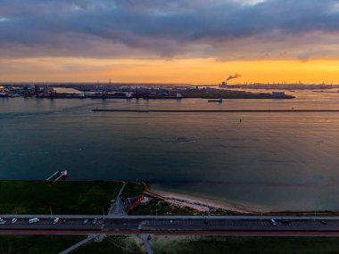 Kıyı sanayi bölgesi yolu, sahil ve çimenli kıyı şeridinin hava günbatımı manzarası dramatik bulutlar altında bacalar, turnalar ve dalgakıranlarla tezat oluşturuyor..