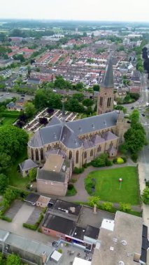 Dik çatısı, kemerli pencereleri ve bayrakları olan Gotik tarzı taş bir kilisenin havadan görünüşü modern gabollu evlerin ve küçük bahçelerin yoğun mahallelerinin yanındaki manzaralı alanlarda sergileniyor..