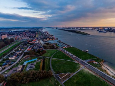 Günbatımı kargo gemileri, vinçler ve endüstriyel rıhtımlardaki nehir limanının havadan görünüşü, yoğun şehir şebekesi ve yeşil alanlar dinamik ticari bir manzarada su yolunu çerçeveliyor..