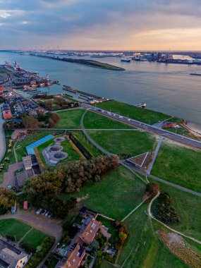 Günbatımı kargo gemileri, vinçler ve endüstriyel rıhtımlardaki nehir limanının havadan görünüşü, yoğun şehir şebekesi ve yeşil alanlar dinamik ticari bir manzarada su yolunu çerçeveliyor..