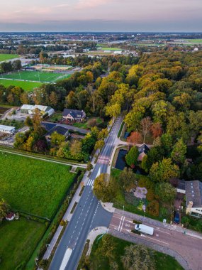 Banliyö manzarası merkez yol, evler, spor sahaları, kanallar ve sonbahar yeşillikleriyle kaplı orman bölgesi, harmanlanmış konut planlaması