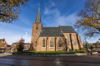Uzun çan kulesi ve saat kulesi ile tarihi tuğla kilise, kemerli pencereler ve kayrak çatı çerçeveli sonbahar ağaçları, plaza ve banklar bu sakin köy sahnesine cazibe ekler açık mavi gökyüzü altında.