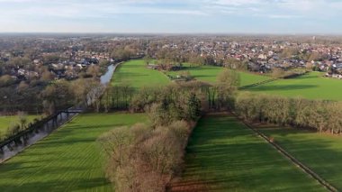 Aerial view of Dutch countryside green fields, sports complex, canal, and quiet town framed by tree lined roads and soft light, blending land use and natural serenity.
