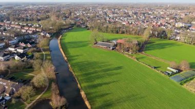 Aerial view of Dutch town dense homes, winding river, and green fields with garden plots blend suburban planning and natural landscape in harmonious layout.