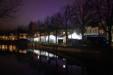 Geceleri sisli Hollanda kanalı çadırları, kilise kulesi ve parlayan yansımalar ağaçların, binaların ve polis minibüsünün karanlık gölgeleriyle kontrastlı sıcak ışıklar..
