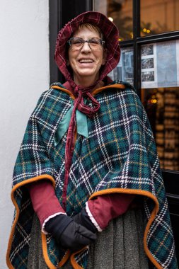Deventer, Overijssel, Hollanda - 14.12.2025: Charles Dickens Festivali 'nde. Ekose kapüşonlu ve pelerinli gülümseyen kadın Victoria dönemi kıyafetleriyle süslenmiş pencerenin önünde duruyor..