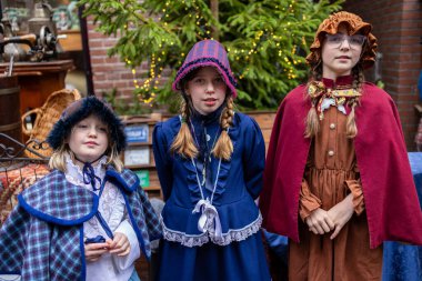Deventer, Overijssel, Hollanda - 14.12.2025: Charles Dickens Festivali 'nde. Kapüşonlu ve pelerinli üç çocuk festival ağacının yanında poz veriyor ve tarihi giysiler içinde klasik aksesuarlar giyiyorlar..