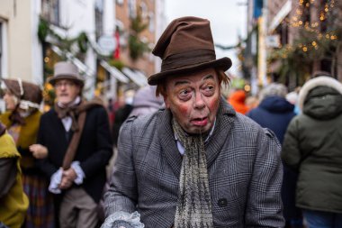 Deventer, Overijssel, Hollanda - 14.12.2025: Charles Dickens Festivali 'nde. Kareli ceketli ve teatral makyajlı bir adam dekore edilmiş Victoria tarzı sokak sahnesinde gösteri yapıyor..