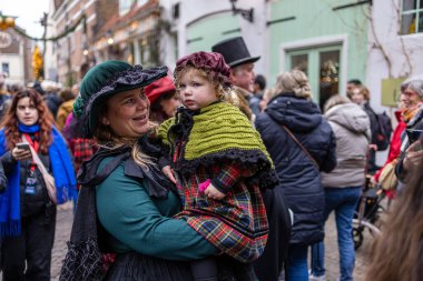Deventer, Overijssel, Hollanda - 14.12.2025: Charles Dickens Festivali 'nde. Yeşil elbiseli kadın, tarihi sokak olaylarında şenlikli kalabalığın ortasında ekose şallı bir çocuk tutuyor..