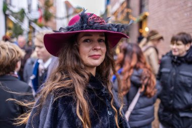 Deventer, Overijssel, Hollanda - 14.12.2025: Charles Dickens Festivali 'nde. Burgonya tüylü şapkalı kadın süslü kış caddesinde şenlikli kalabalıkta göze çarpıyor..