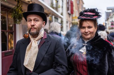 Deventer, Overijssel, Hollanda - 14.12.2025: Charles Dickens Festivali 'nde. Süslü şapkalı ve pelerinli Viktoryalı çift, festival sokaklarında toplanan duman ve kalabalığın arasında poz veriyor..