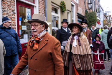 Deventer, Overijssel, Hollanda - 14.12.2025: Charles Dickens Festivali 'nde. Bronzlaşmış silindir şapkalı ve çizgili kravatlı adam kaldırım taşı sokağında çelenklerle Victoria dönemi geçit törenine öncülük ediyor..