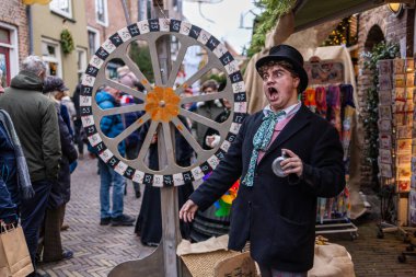 Deventer, Overijssel, Hollanda - 14.12.2025: Charles Dickens Festivali 'nde. Şenlikli kaldırım taşı pazarında silindir şapkalı ve eşarplı bir sanatçı numaralandırılmış dönen çarkın yanında jestler yapıyor..