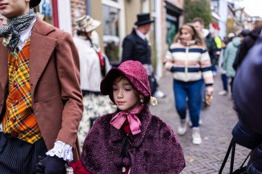 Deventer, Overijssel, Hollanda - 14.12.2025: Charles Dickens Festivali 'nde. Burgundy şapkalı ve pelerinli bir çocuk festivalde dantel kelepçeli bir yetişkinle el ele yürüyor..