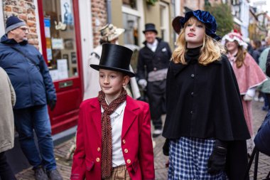 Deventer, Overijssel, Hollanda - 14.12.2025: Charles Dickens Festivali 'nde. Viktorya dönemi elbiseli iki genç festival caddesinde başlıkları, pelerinleri ve ekose pantolonları ile poz veriyorlar..