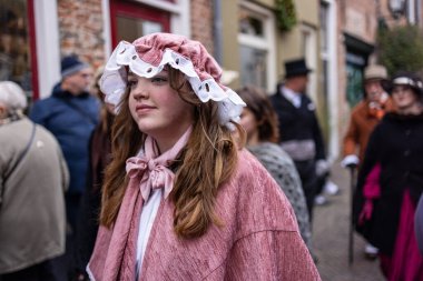 Deventer, Overijssel, Hollanda - 14.12.2025: Charles Dickens Festivali 'nde. Pembe şapkalı ve pelerinli çocuk eski binalarla birlikte kaldırım taşı caddesinde Victoria dönemi kalabalığının ortasında duruyor..
