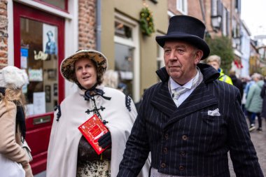 Deventer, Overijssel, Hollanda - 14.12.2025: Charles Dickens Festivali 'nde. Sokak kutlaması sırasında çelenk desenli dükkanın önünden kitap gezintisi yapan Viktoryalı çift..
