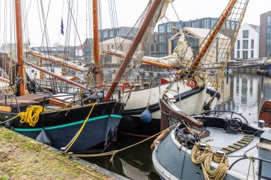 Geleneksel yelkenli tekneler, ahşap direkleri ve sarmalanmış donanımlarıyla sakin bir limanda dinleniyorlar. Klasik formları, rıhtım boyunca uzanan modern ve gösterişli binalar..