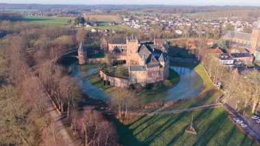 Hendek ve kuleleri olan kırmızı tuğlalı ortaçağ şatosunun havadan görünüşü. Yapraksız ağaçlar, kilise kulesi ve uzun kış gölgeleri olan yarı kırsal bir kasaba..
