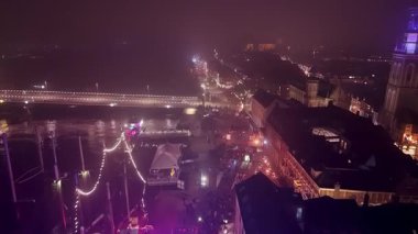 Aydınlatılmış köprü, kenetlenmiş tekneler, parıldayan saat kulesi ve hareketli sokak panayırı ile gece hava manzarası.