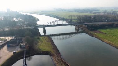 Kırsal alanda kıvrımlı nehrin üzerindeki kemerli köprünün havadan görünüşü. Araçlar takviyeli bankaların üzerinden geçiyor, yeşil tarlalar, ağaçlar ve karışık kullanım bölgeleriyle çevrili..