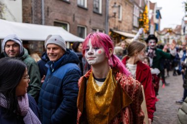Deventer, Overijssel, Hollanda - 14.12.2025: Charles Dickens Festivali 'nde. Pembe saçlı ve kostümlü yüz boyalı kişi kalabalık ve ışıklarla geçit törenine öncülük ediyor..