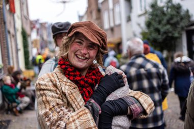 Deventer, Overijssel, Hollanda - 14.12.2025: Charles Dickens Festivali 'nde. Eski moda şapkalı ve eşarplı gülümseyen insan kostümlü kalabalıkla dolu neşeli bir caddeye sarılmış..